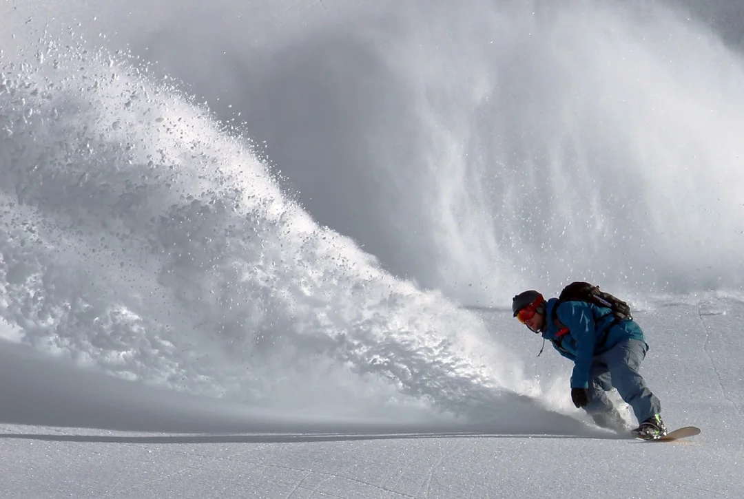 A skier dropping into a steep, untracked powder field in the backcountry, mountains and blue sky behind them, wearing a full touring setup with splitboard visible on a nearby slope